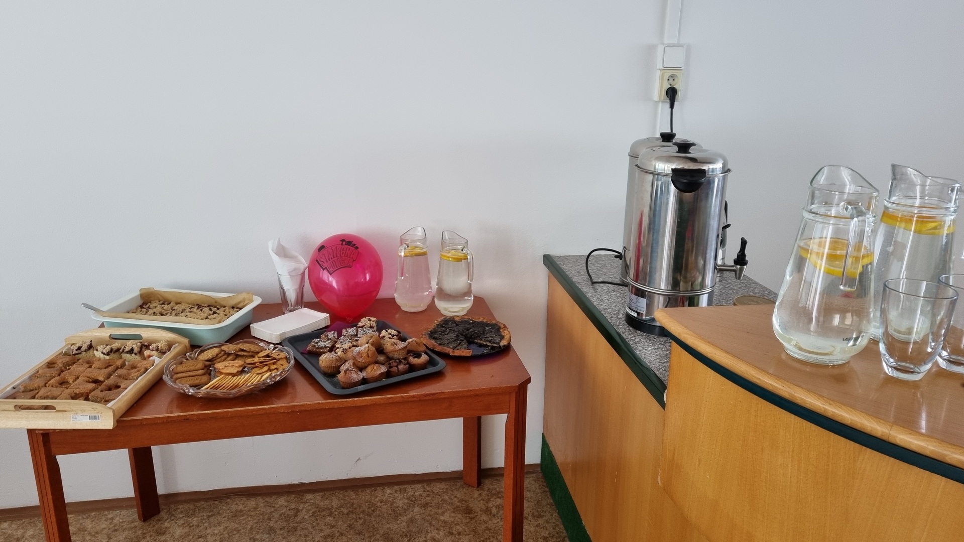 pohled na stoly s občerstvením - tácy s buchtami, džbánky s vodou a citronem a várnice s horkou vodou pro přípravu kávy a čaje
