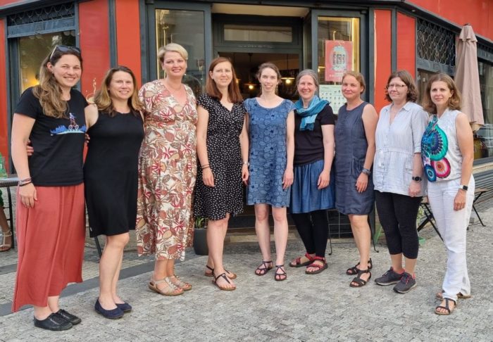 9 žen stojí vedle sebe v půlkruhu před otevřenými dveřmi do kavárny