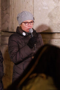 Před zdí budovy stojí žena v zimní bundě, čepici a rukavicích a hovoří do mikrofonu.